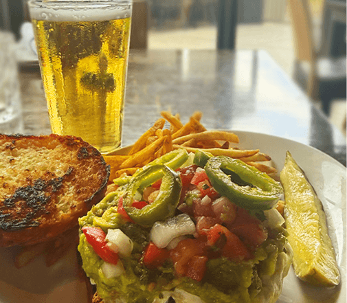 Open-face Sonoran burger topped with guacamole, pico de gallo, and sliced jalapeños, served with fries, a pickle, and a pint of draft beer.