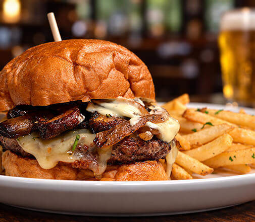 A thick juicy burger stacked with melted Swiss cheese and sautéed mushrooms on a toasted brioche bun, served with golden french fries and a glass of beer.
